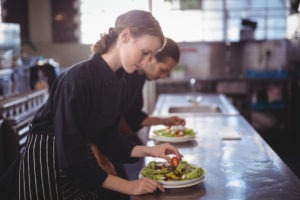 Restaurant staff prepare salad on a commercial kitchen island.