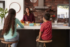 Kids eat breakfast while mother prepares school lunch on a kitchen island.